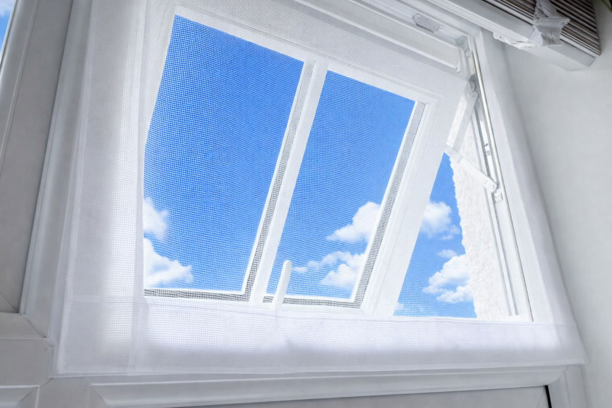 White window mesh screen installed on a white window frame, showing a bright blue sky with clouds outside.