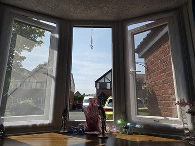 A three-bay bay window with white frames installed on a curved wall, showing a residential street scene outside with houses, parked cars, and a brick wall. The interior window ledge has a wooden surface with small decorative items in front of the windows.