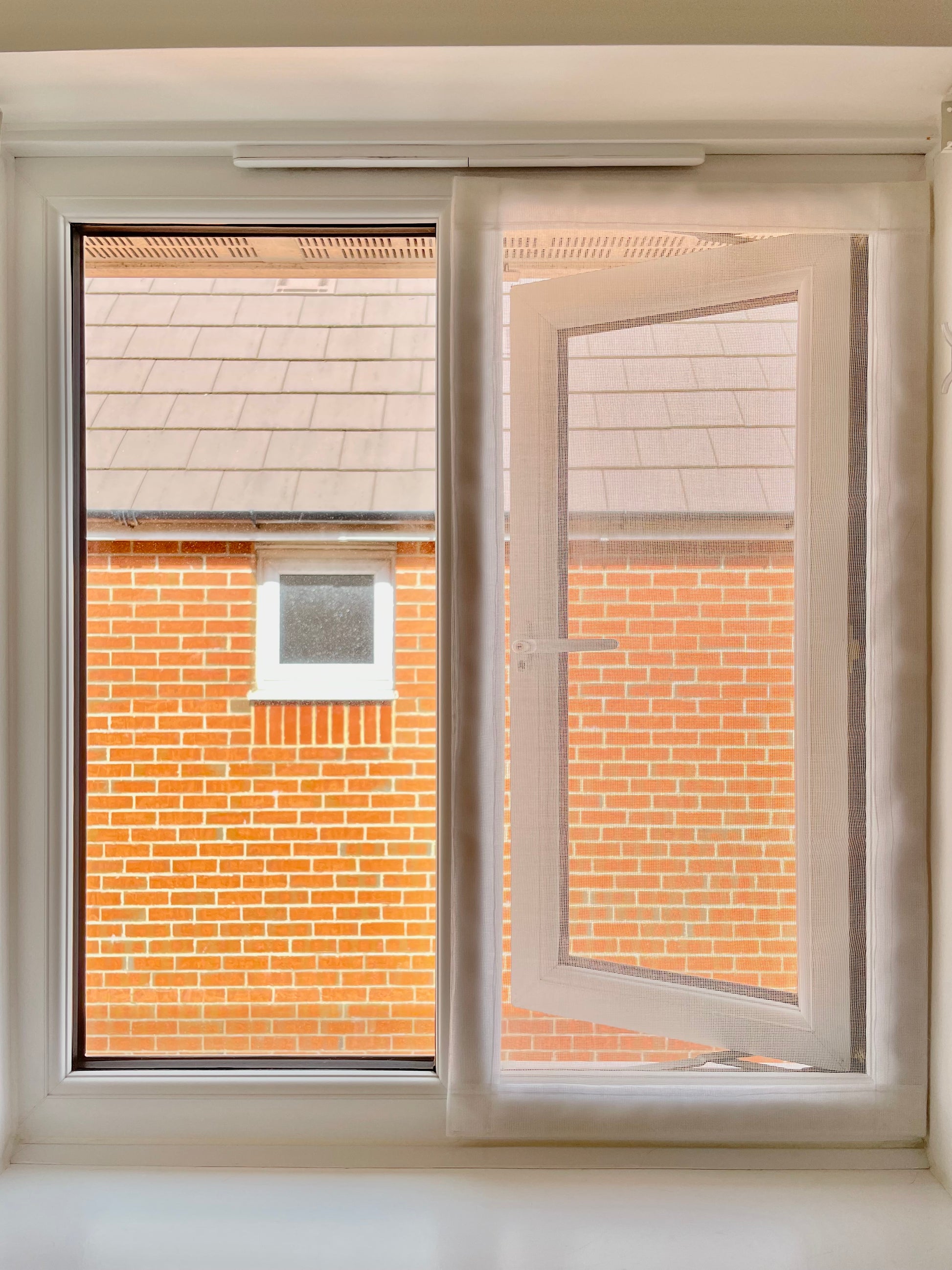 A white uPVC window with one panel open, showing a brick building exterior through the glass, with a white window screen mesh attached.